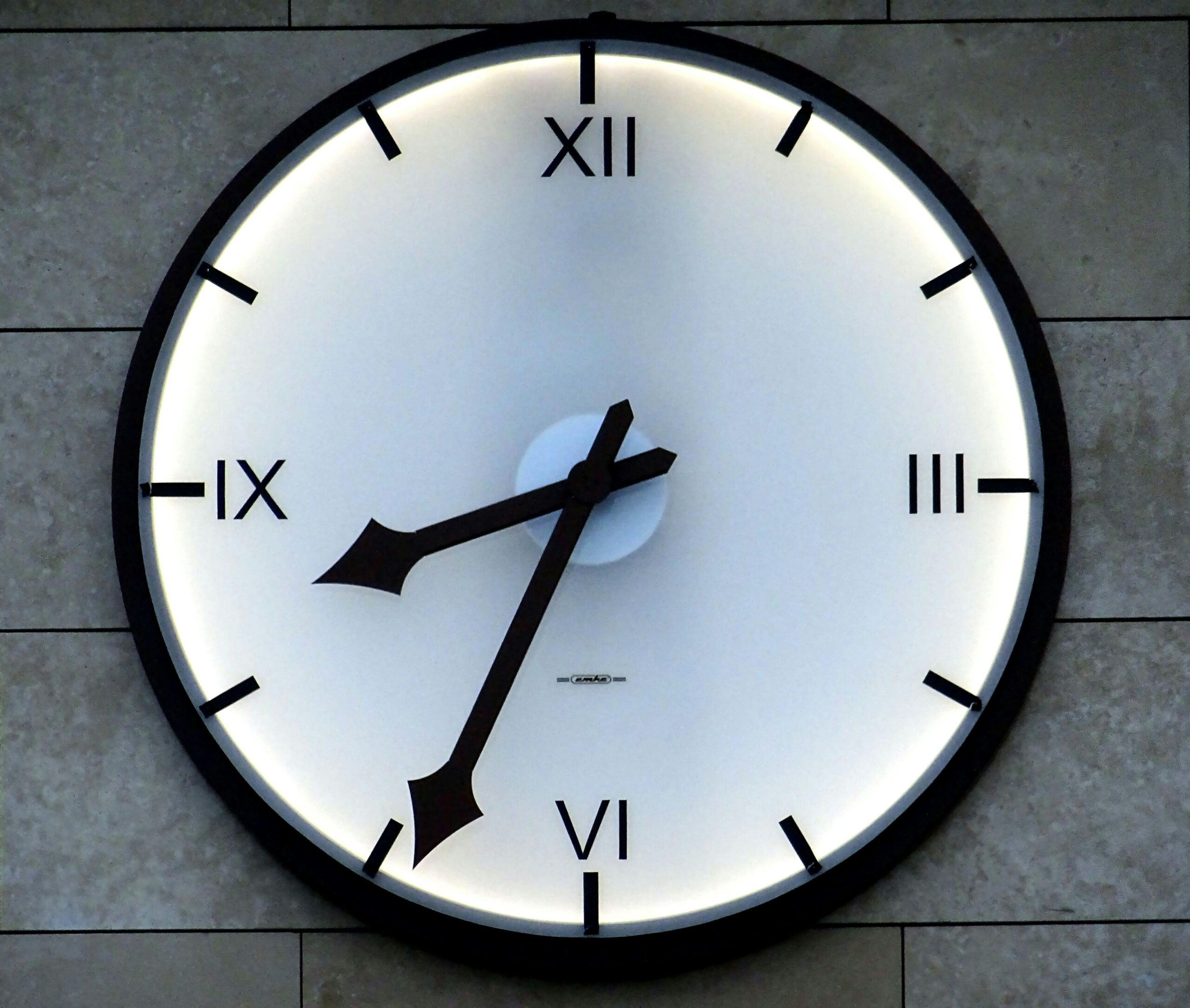 Close-up of a modern wall clock with Roman numerals and backlit design, emphasizing timekeeping precision.